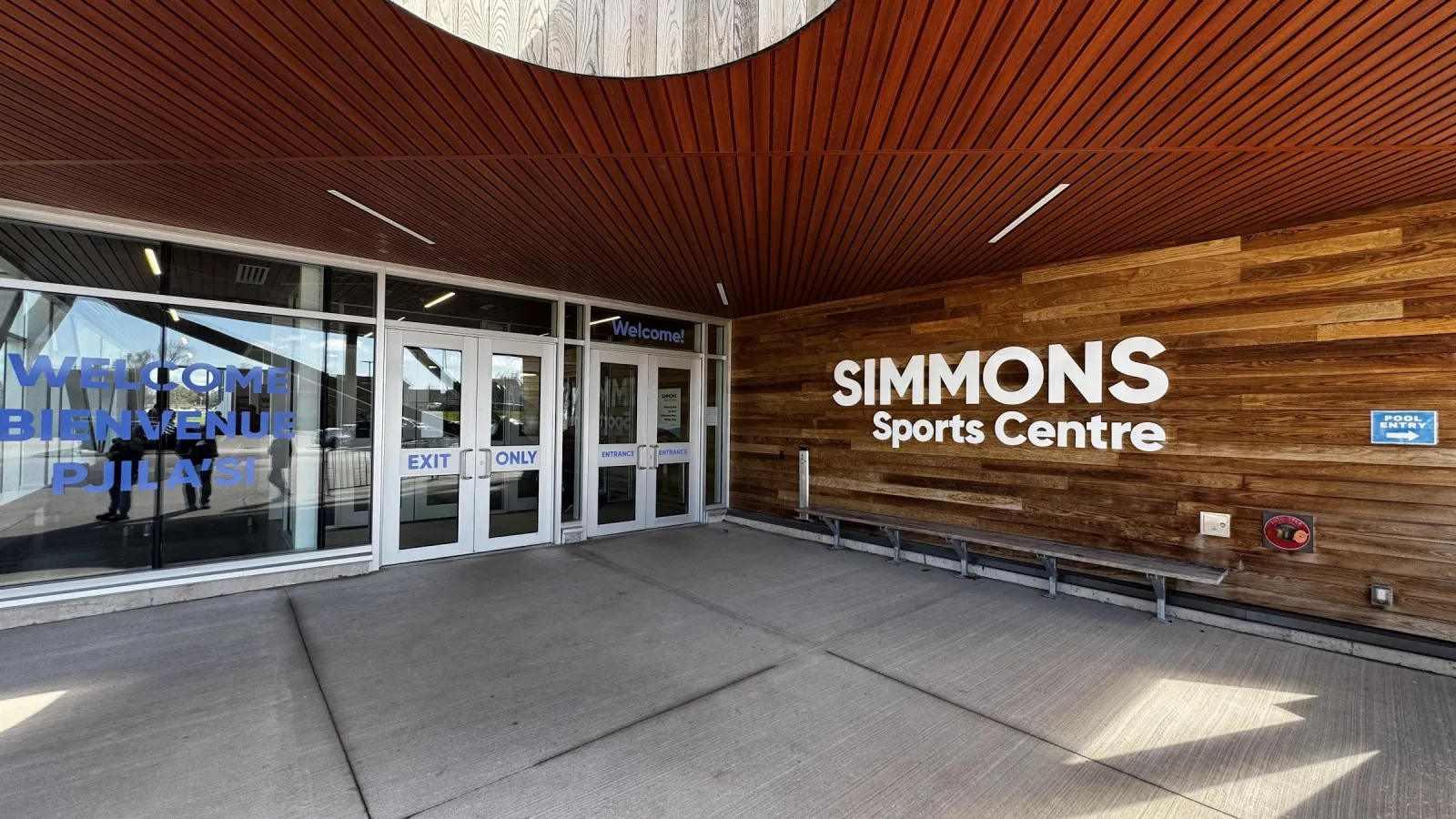 La entrada de vidrio con Simmons Sports Centre rotulado en un muro de listones de madera bajo un plafón de madera, en un día soleado. — Simmons Sports Centre