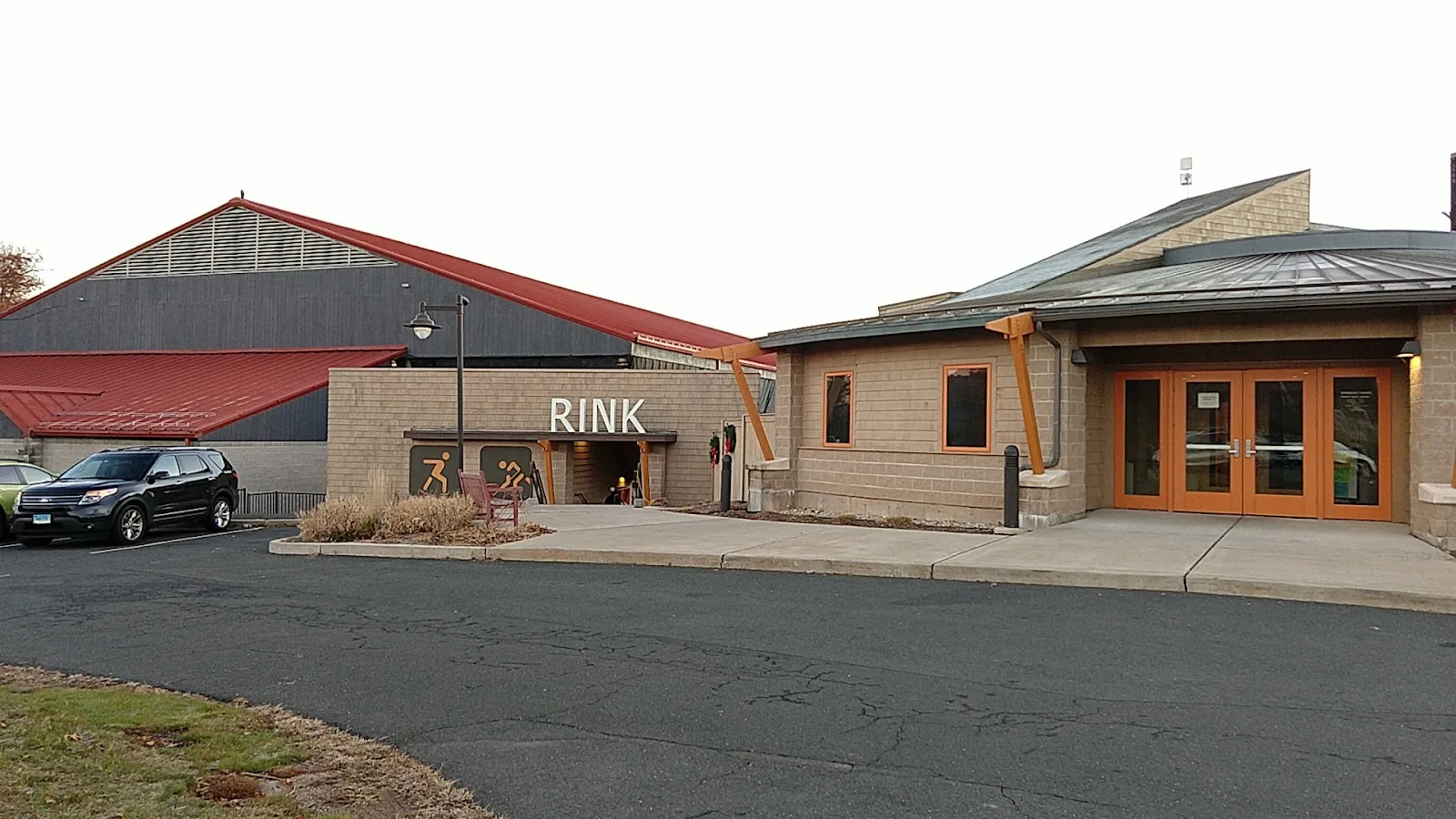 The rink building's entrance with a red-roofed arena behind it, the sign RINK above the door and the stone-and-timber lobby beside the car park. — Simsbury Farms Ice Rink
