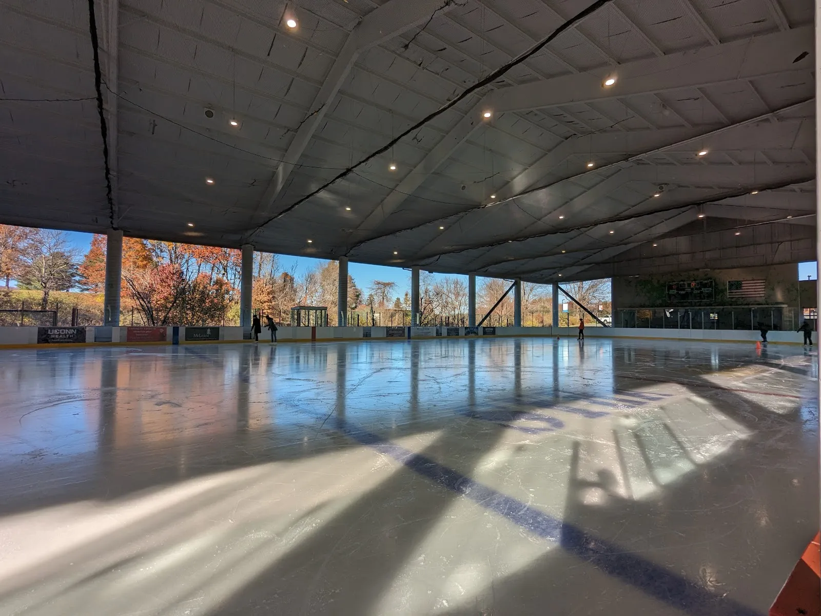 The covered, open-sided rink with its ice reflecting the autumn trees beyond the columns and a few skaters at the far end; a blue line crosses the ice. — Simsbury Farms Ice Rink