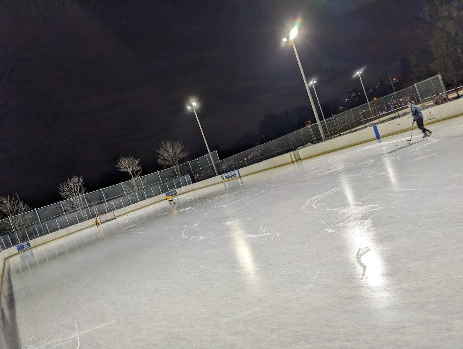 Pista al aire libre iluminada con reflectores, con bardas y cercas, en Sir Adam Beck Park; la superficie de hielo en primer plano y un patinador a lo lejos — Sir Adam Beck Park Outdoor Ice Rink