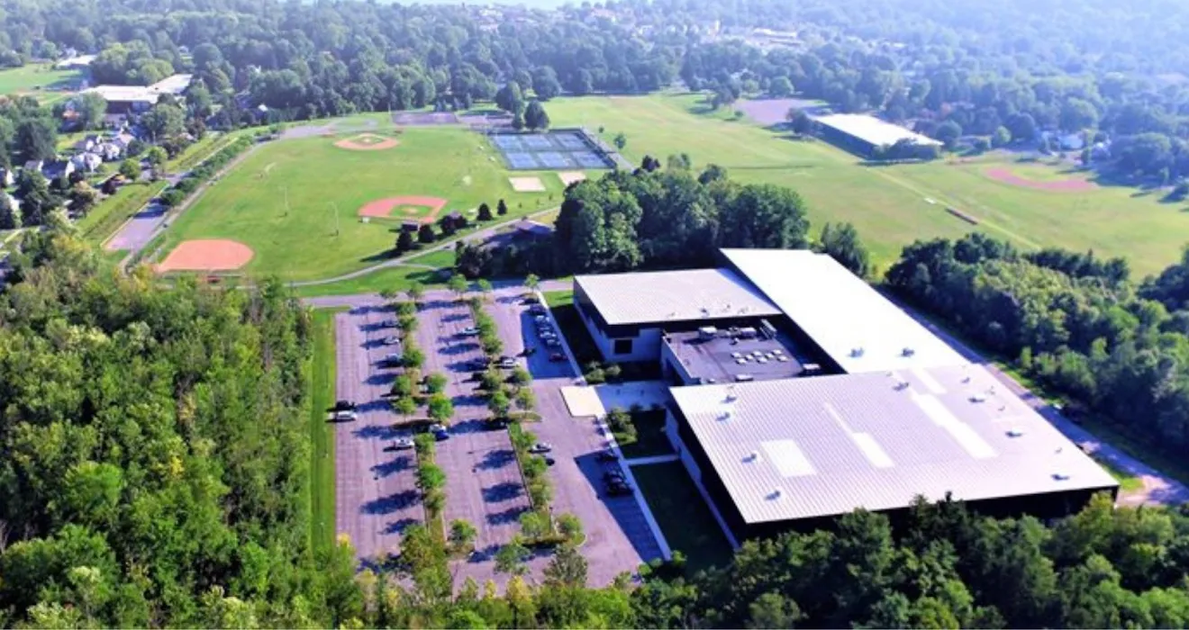 Vista aérea del Skaneateles Community Center, el gran edificio de techo plano junto a su estacionamiento bordeado de árboles, con campos de béisbol detrás — Skaneateles Community Center