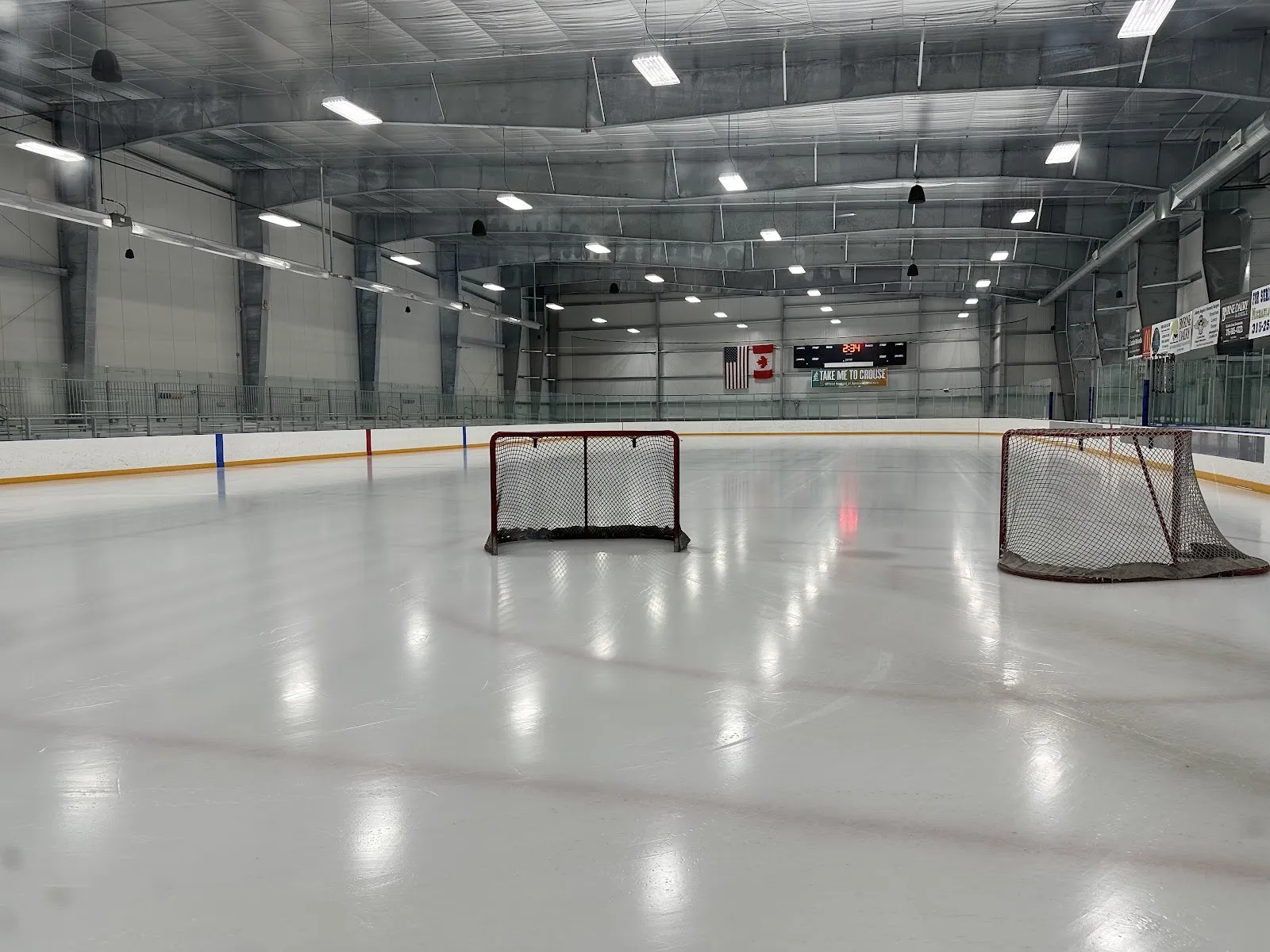 Una pista vacía con dos porterías de hockey en el hielo, un marcador entre las banderas de Estados Unidos y Canadá al fondo y cristal a ambos lados. — Skaneateles Community Center