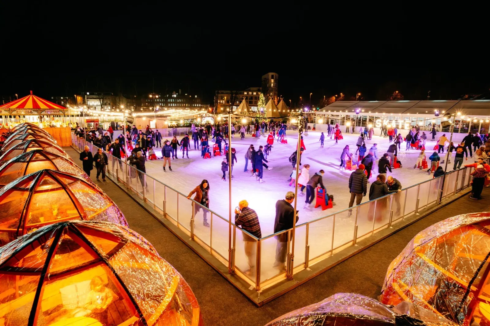The Skate Cambridge ice rink from above at night in the 2024 season: the whole rink behind its barrier with skaters spread across the ice, lit igloo huts in the foreground. Set-up for 2026 may differ. — SKATE Cambridge