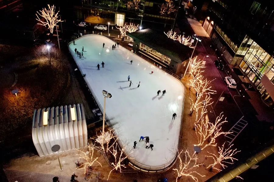 Aerial view at night of the Canal District Kendall rink, a pale oval of ice ringed by lights between the glass towers. — Skate @ Canal District Kendall