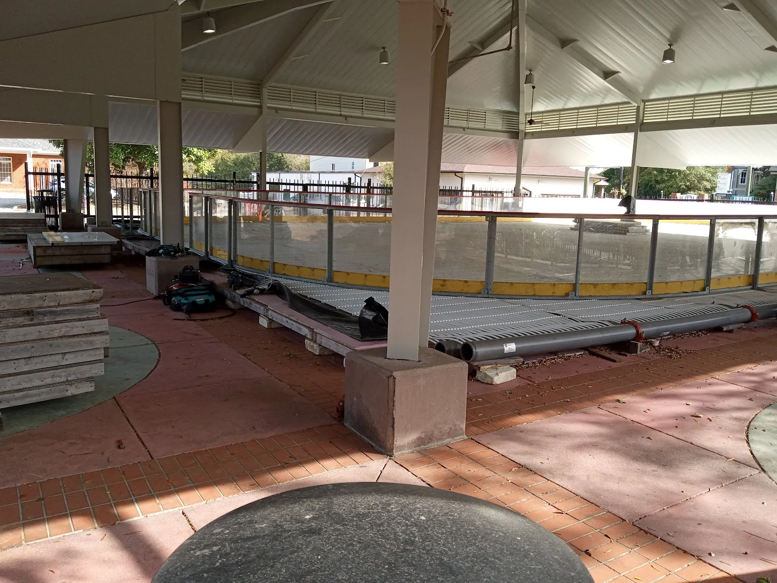 The open-sided roofed pavilion at Olde Town Conyers' Celebration Park in daylight, with the empty rink under the roof, rail posts and a covered walkway around it. — Skate Conyers (Ice Days)