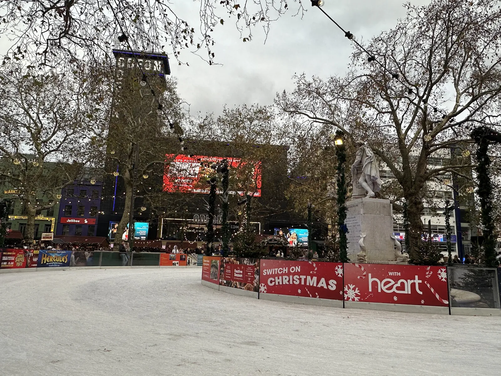 Leicester Square in November 2025 with the rink curving round the Shakespeare statue, bare plane trees and the Odeon cinema behind. — Skate Leicester Square