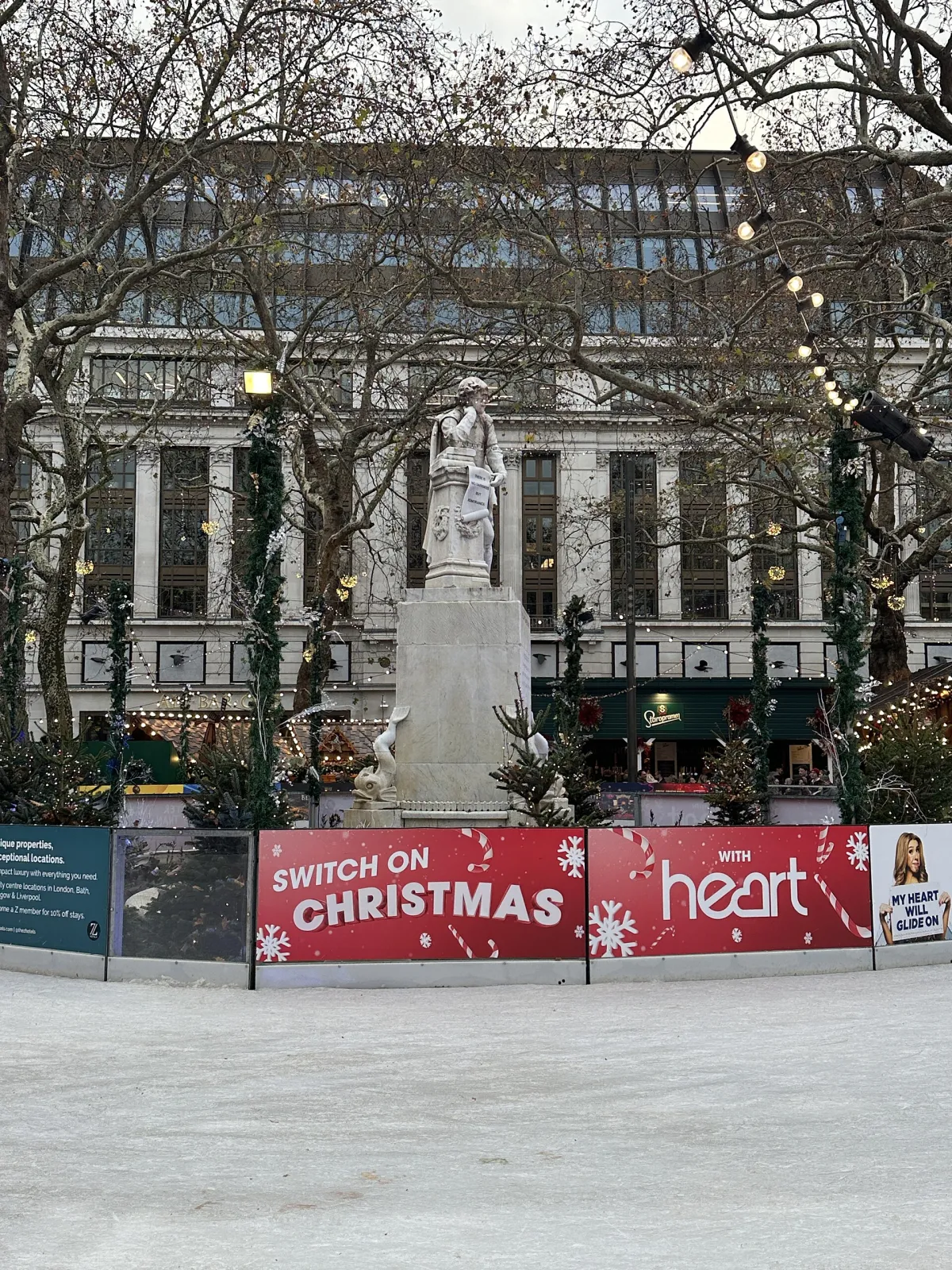 The empty ice in front of the Shakespeare statue, ringed by the rink barrier with Christmas sponsor boards and a building facade behind. — Skate Leicester Square