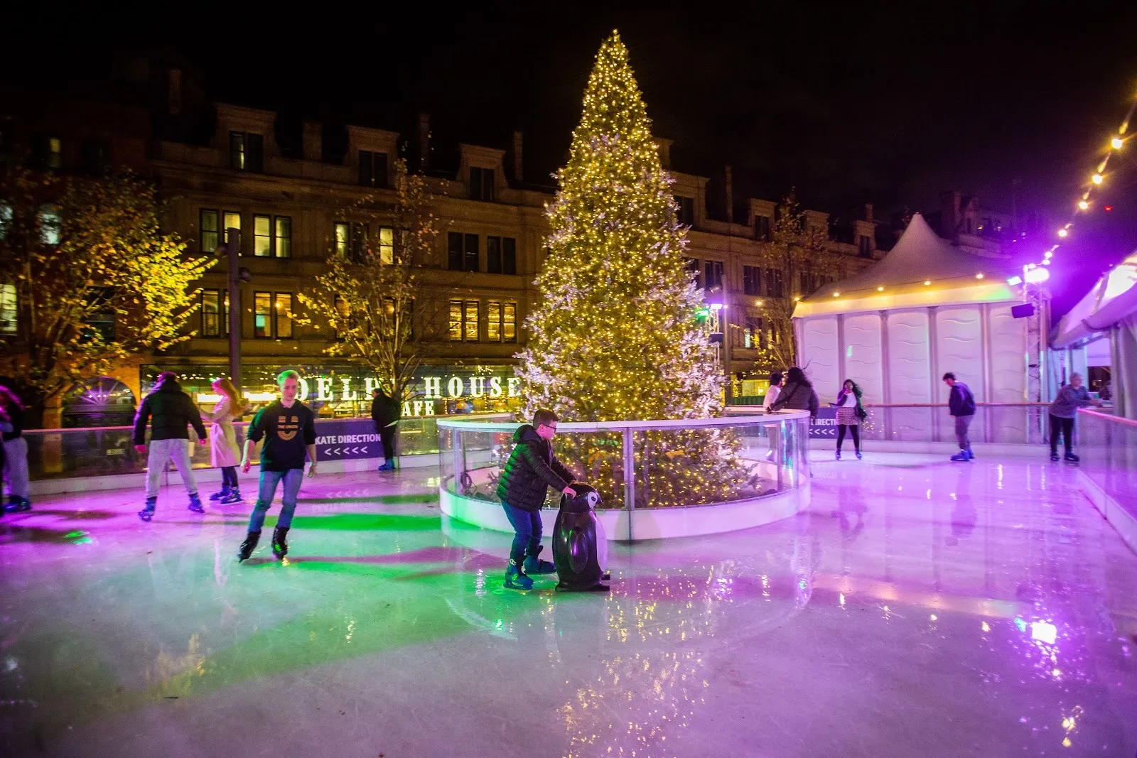 The Skate Manchester rink in Cathedral Gardens at night, skaters on the lit ice beside a tall Christmas tree and the market chalets. — Skate Manchester