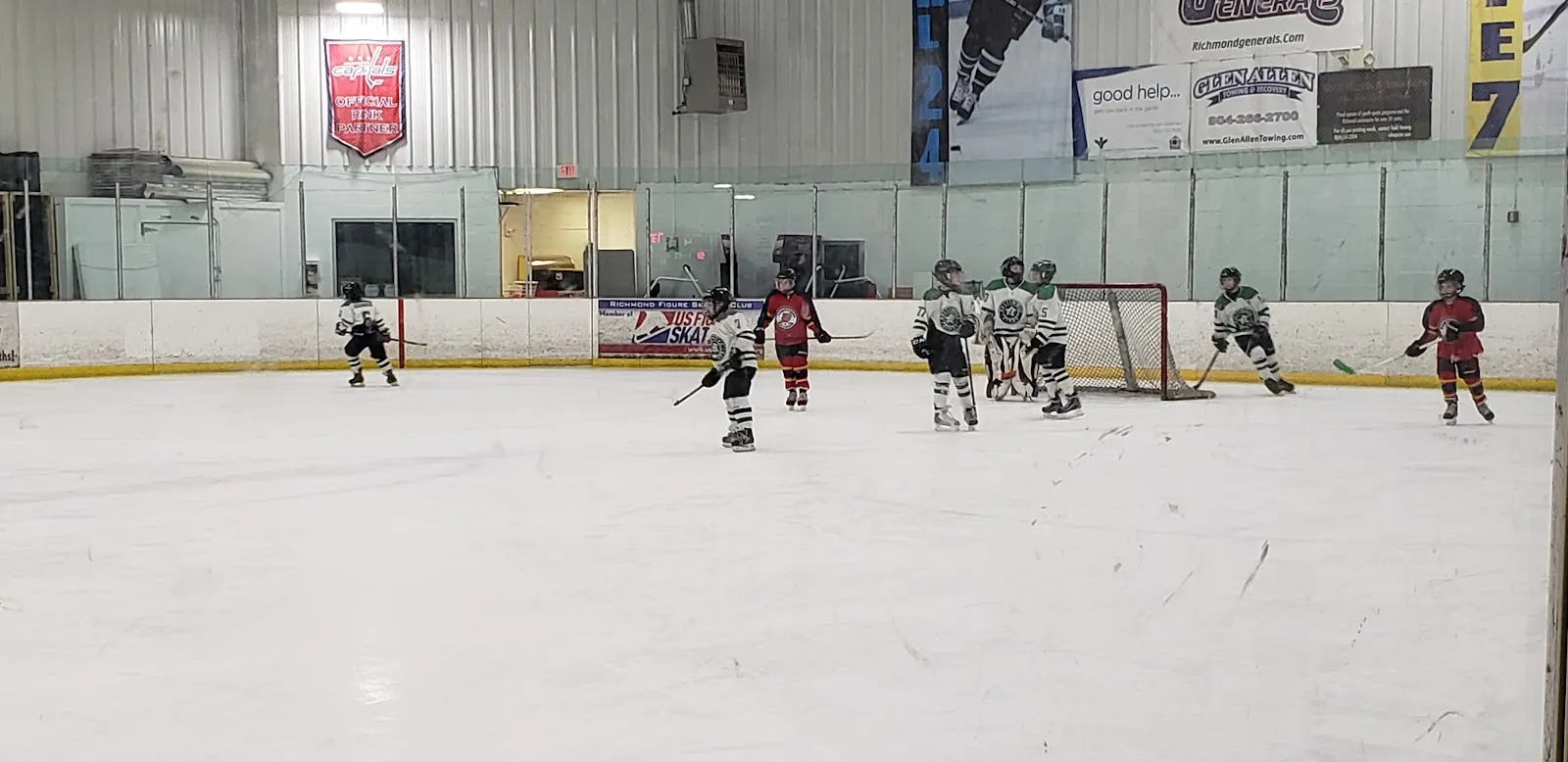 A youth hockey practice at the far end of the sheet, small players across the ice, with banners and flags along the end wall and the glass in front. — SkateNation Plus (Glen Allen)