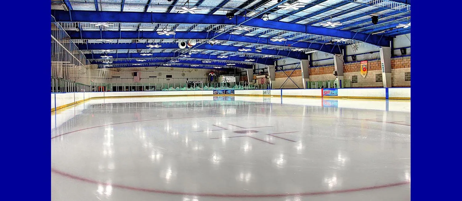 Pista vacía de tamaño NHL en Skatetown, vista amplia hacia las vallas del fondo con el techo de armaduras azules, banderas junto a la pista y una bandera de Estados Unidos visibles. — Skatetown Ice Arena