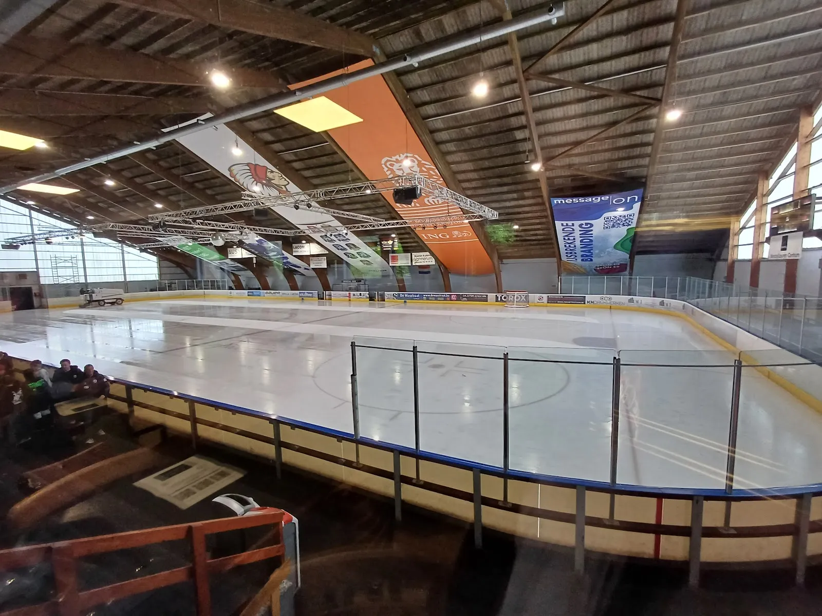 The empty ice sheet at Skateworld in Leuven seen over the boards from the stand, under the timber roof with hanging banners — Skateworld