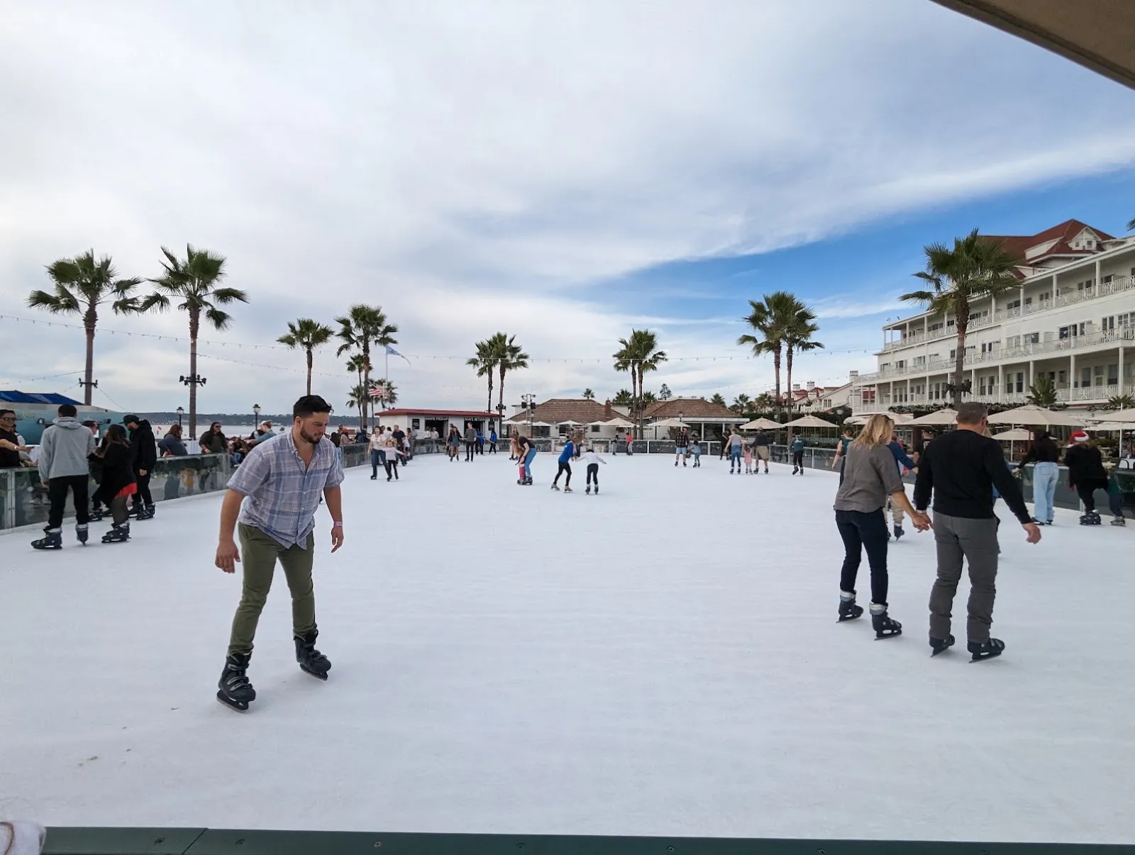 The rink surface in daylight with a few skaters, palm trees along the far edge and the hotel's white buildings behind. — Skating by the Sea (Hotel del Coronado)