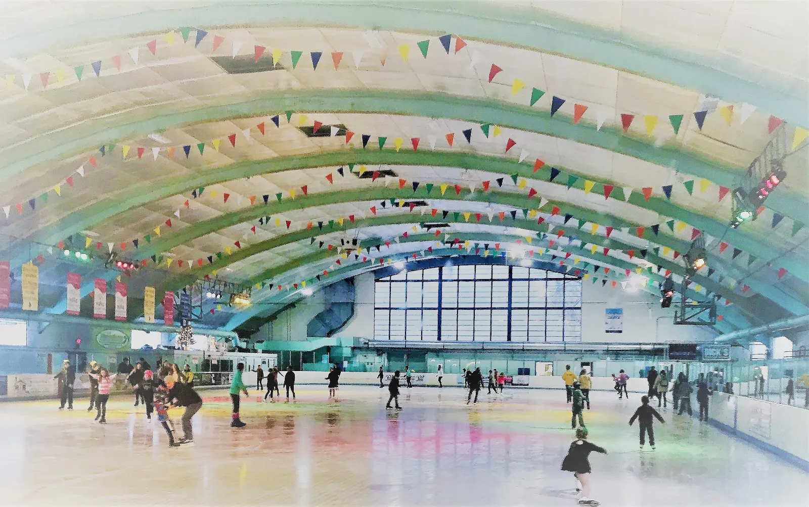 Interior de la pista: un techo de vigas de madera de bóveda de cañón con banderines de colores sobre toda la pista, patinadores del público en el hielo y un alto muro de ventanas en el extremo opuesto. — Skating Club of Wilmington