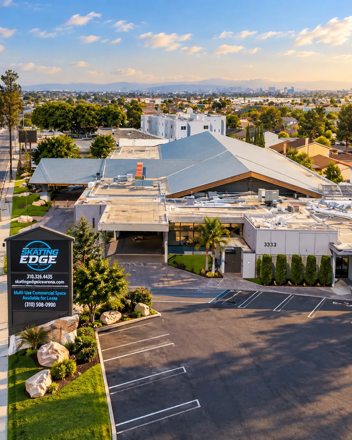Aerial drone view of the Skating Edge building's distinctive peaked roof and front entrance, with the illuminated monument sign reading SKATING EDGE and street address 3333 visible in the parking lot. — Skating Edge Ice Arena