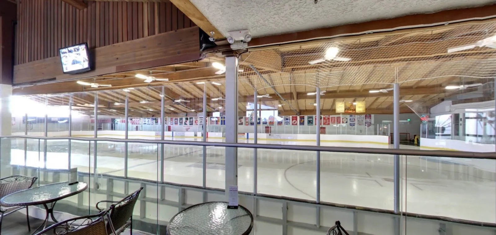 The Skating Edge rink from the upstairs lounge, empty ice under a timber roof hung with team pennants and flags — Skating Edge Ice Arena