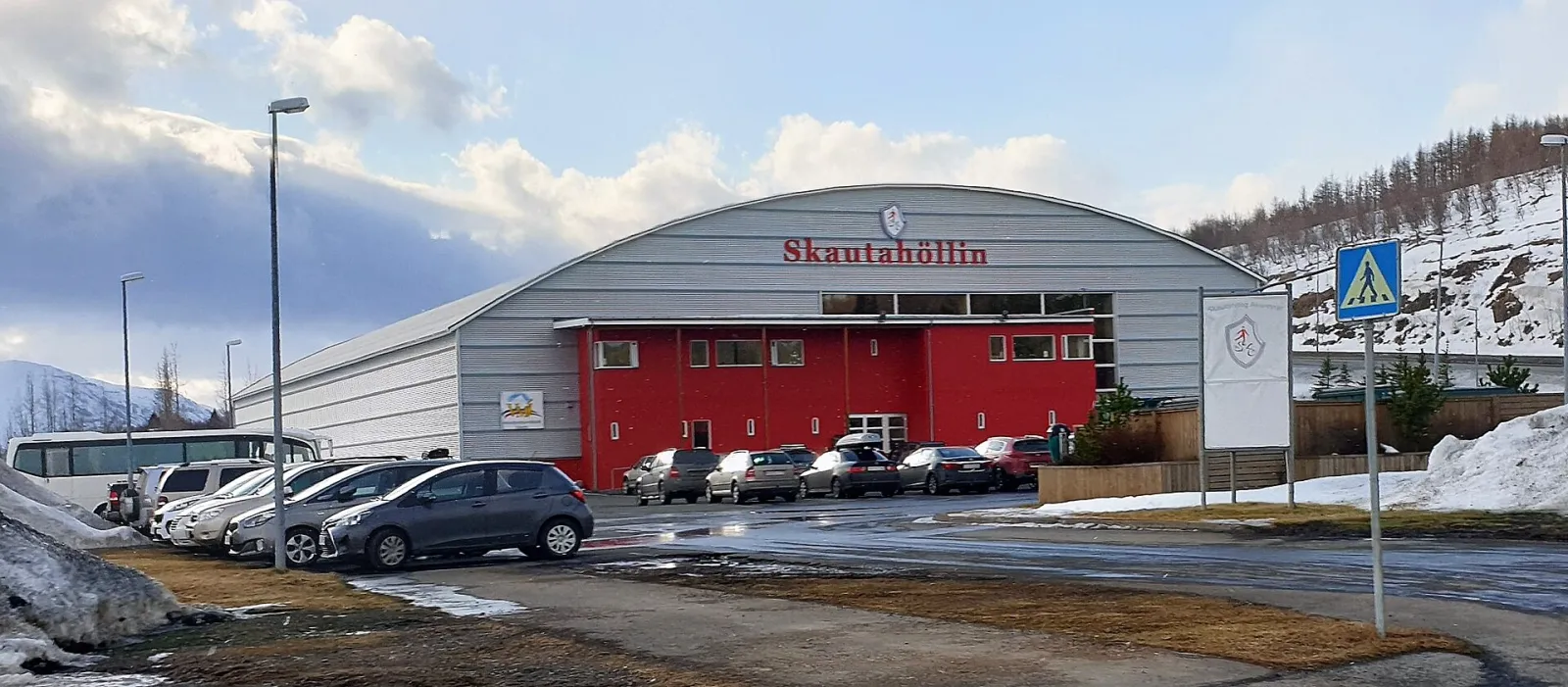 The grey barrel-roofed Skautahöllin á Akureyri with its red entrance wing and the car park in front, seen from the street in winter — Skautahöllin á Akureyri