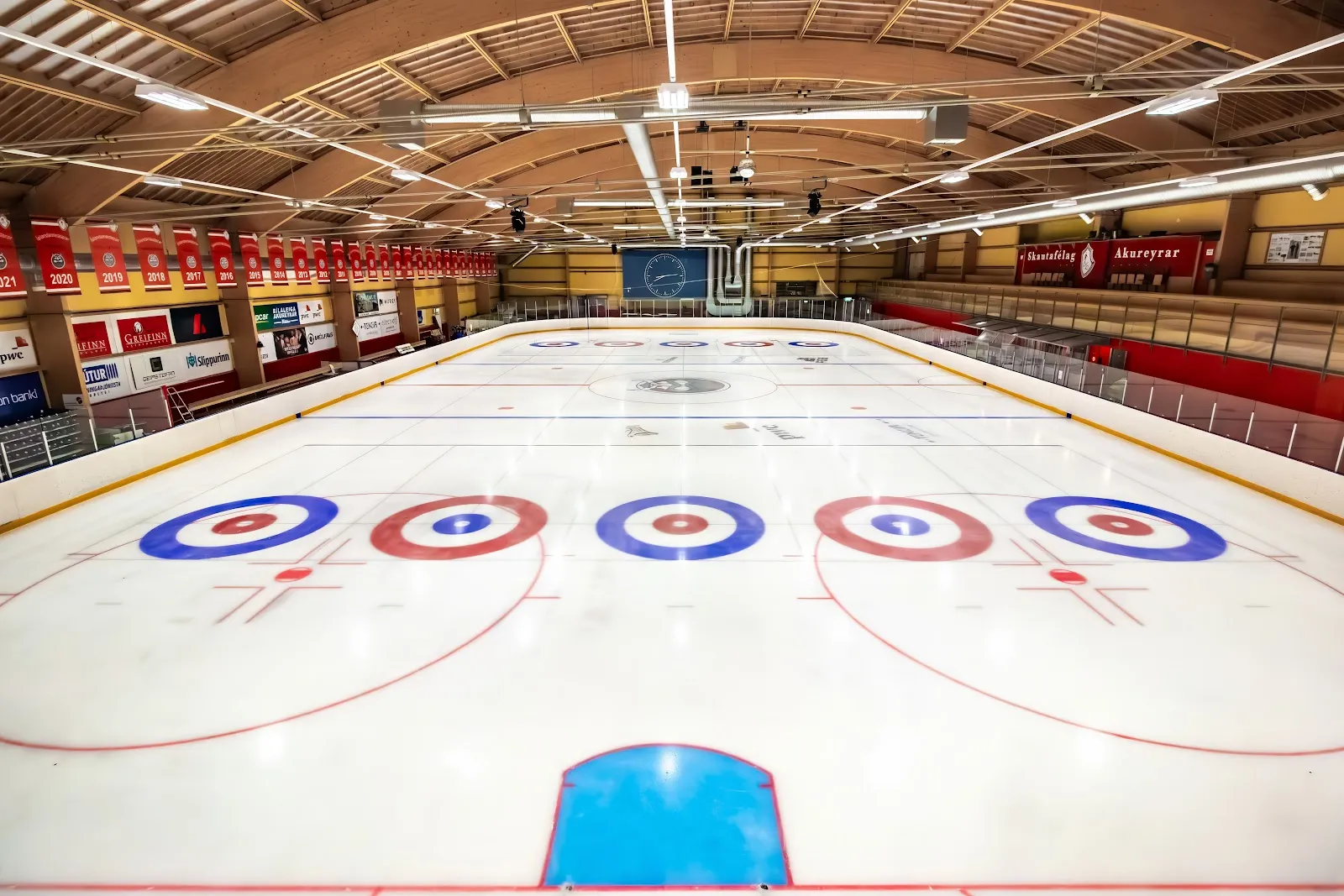 The empty indoor ice sheet seen from one end, with red boards, a barrel-vaulted timber roof and painted circles on the ice — Skautahöllin á Akureyri
