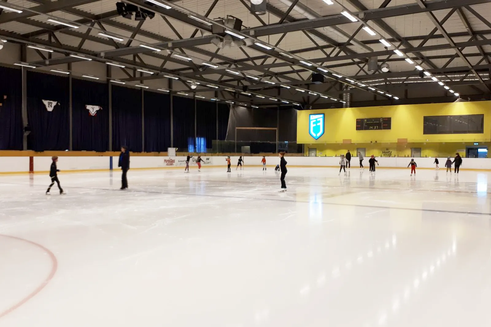 Skaters spread across the ice at Egilshöll under a steel-truss roof, with the yellow end wall, scoreboard and low boards behind them. — Skautasvell Egilshöll (Reykjavík)