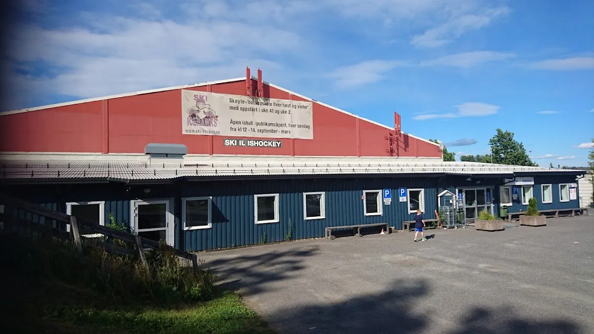 Ski ishall med rødt tak sett over parkeringsplassen, med klubbbanneret på frontgavlen. — Ski Ishall