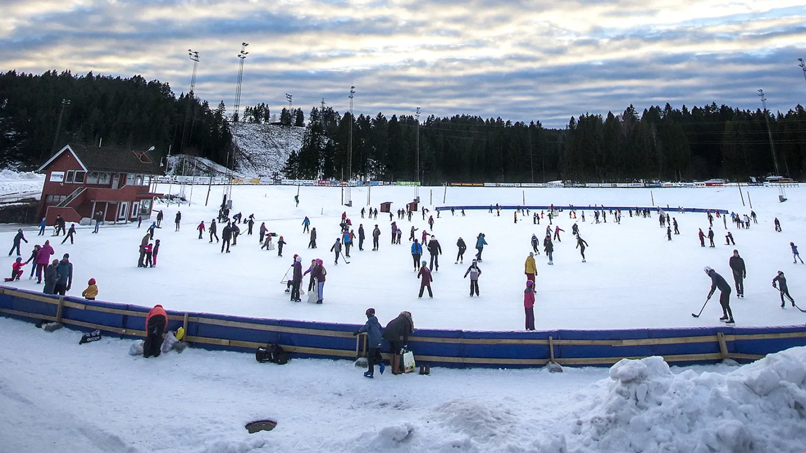 The outdoor skating rink at Skien Fritidspark in winter, a wide open ice surface edged by blue boards, with skaters across it. — Skien Fritidspark skøytebane