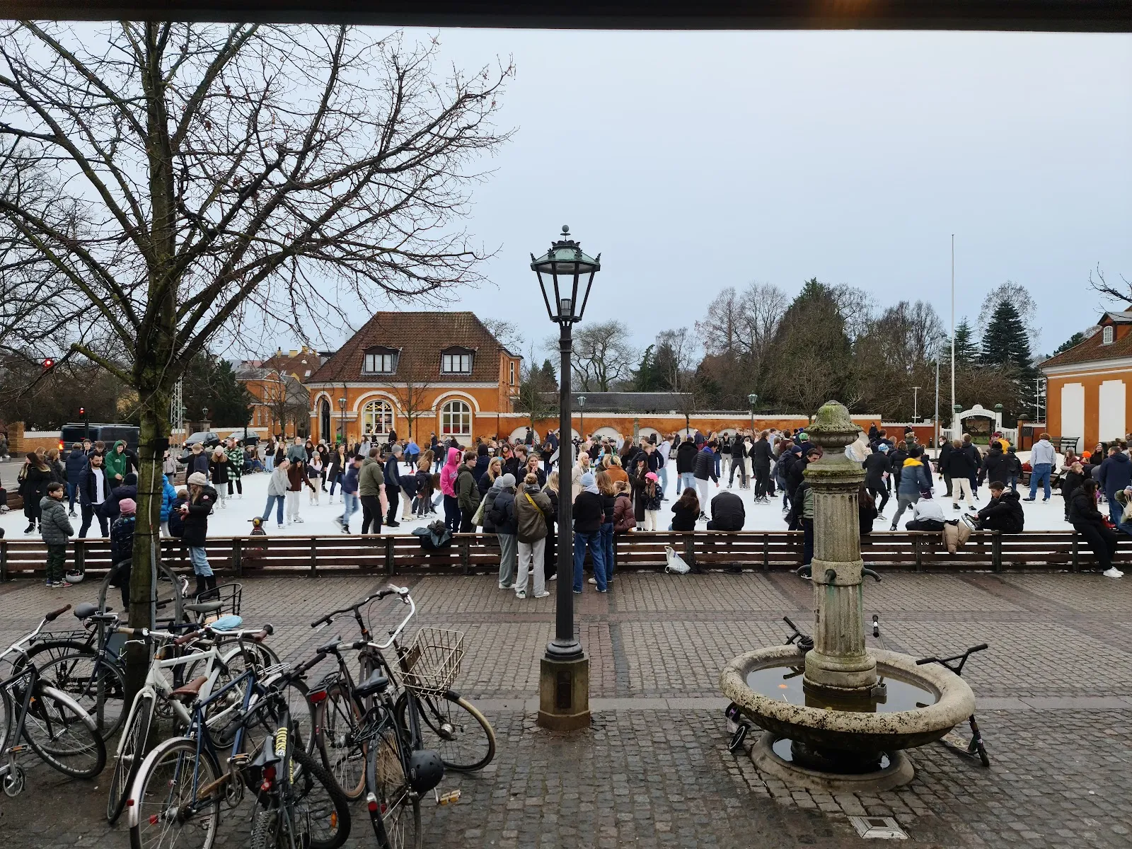 Skøjtebanen på Frederiksberg Runddel set hen over den brostensbelagte plads i skumringen, med de orange portbygninger bag skøjteløberne og en lygtepæl og et springvand foran. — Skøjtebanen på Frederiksberg Runddel