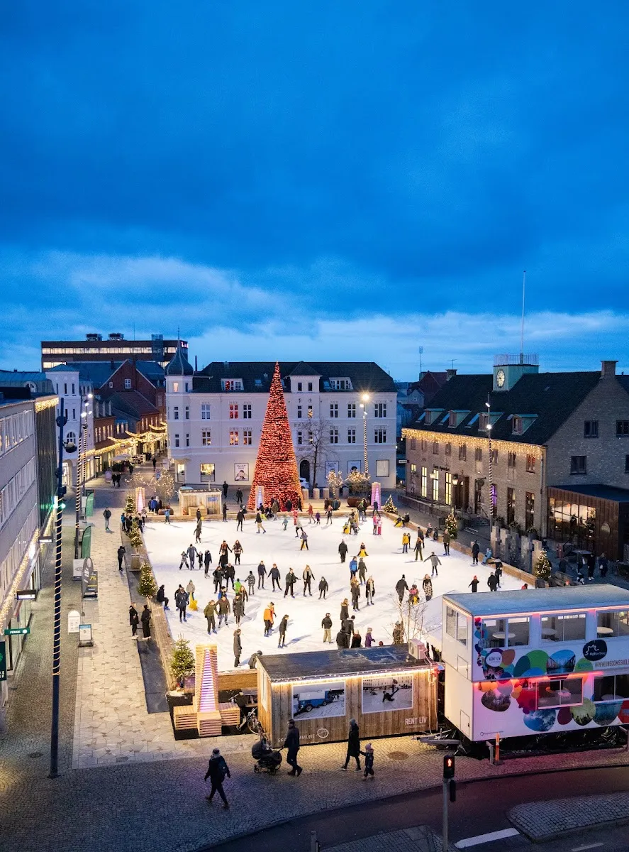 The Skive skating rink on Posthustorvet at dusk, seen from above with the lit Christmas tree, the skate-hire huts and skaters across the ice in the town square. — Skøjtebanen på Posthustorvet