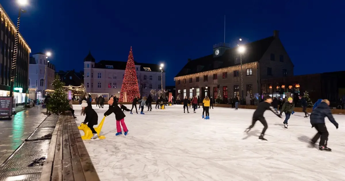 Skaters on the outdoor Skive rink at night, the town square with a lit Christmas tree and old buildings behind. — Skøjtebanen på Posthustorvet