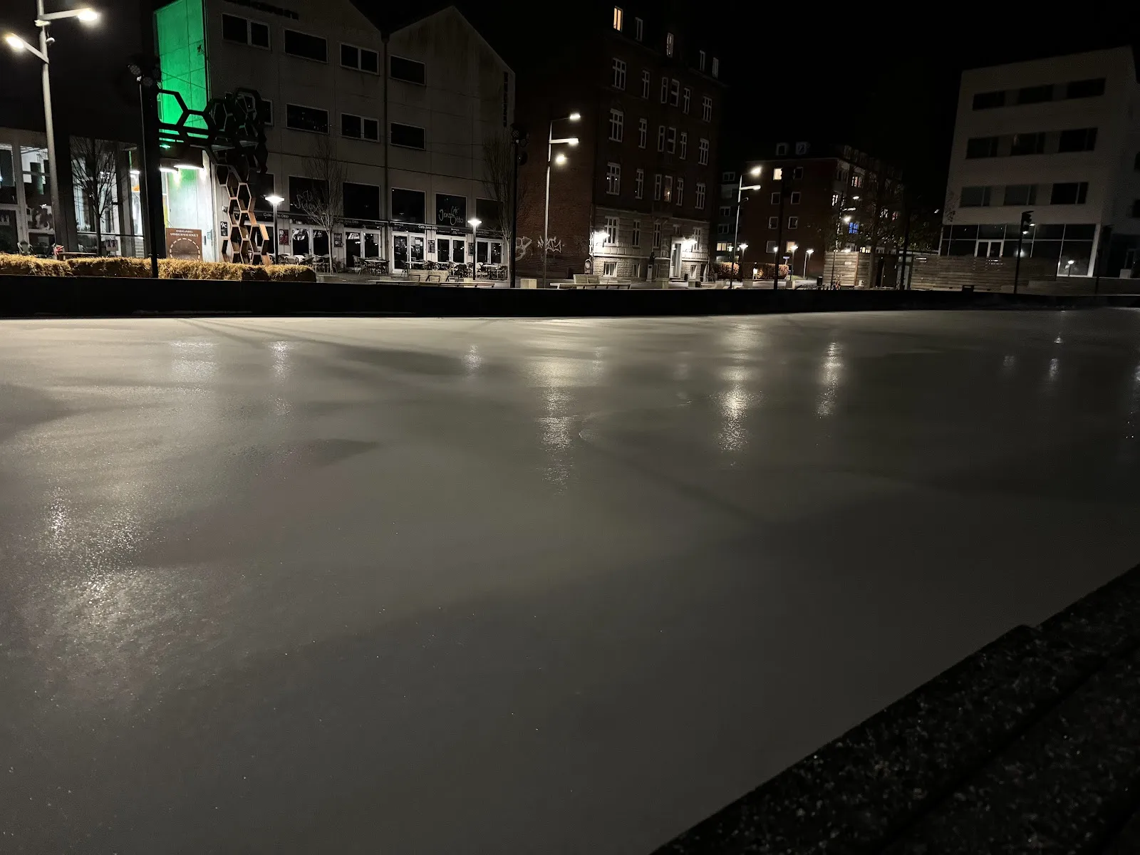 The outdoor rink's smooth ice at night in the town square, with lit apartment and office buildings along the far side. — Randers Skøjtebane
