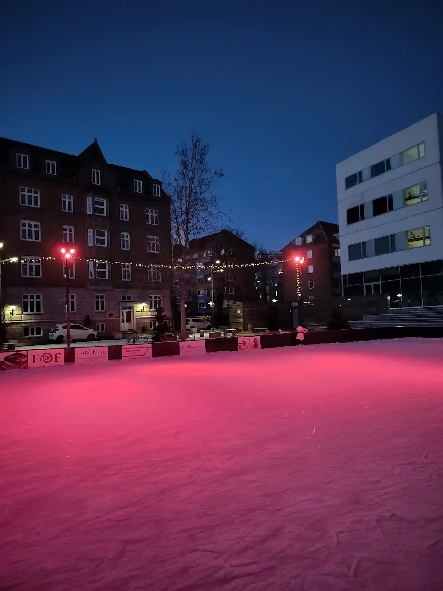 The rink at dusk with its ice lit pink, strings of lights along the boards and brick and white buildings around the square. — Randers Skøjtebane