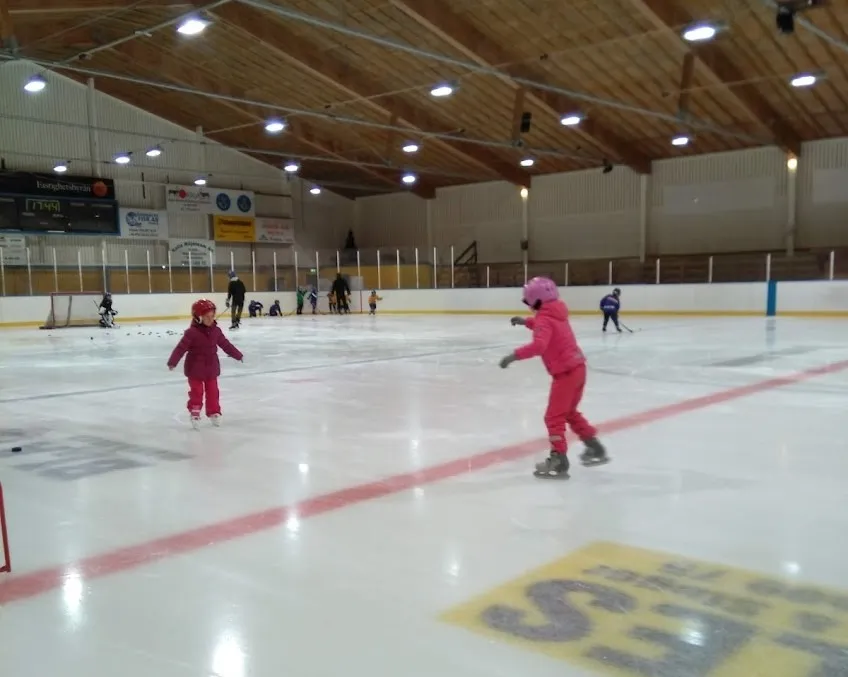 Two children skating on the ice inside Skogshallen ishall in Överkalix, with the timber roof and rink boards behind – Skogshallen (Överkalix ishall)