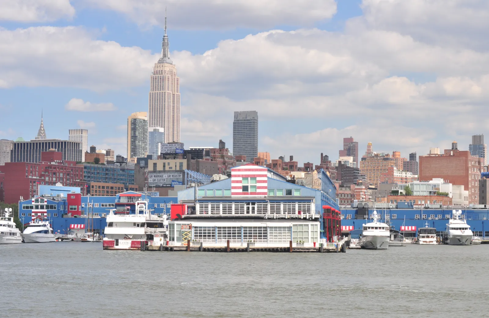 Pier 61, home to Sky Rink, seen from the Hudson River: the blue pier building with its red-and-white-striped roof gable and rows of windows, docked boats along the pier, and the Midtown Manhattan skyline behind. — Sky Rink at Chelsea Piers