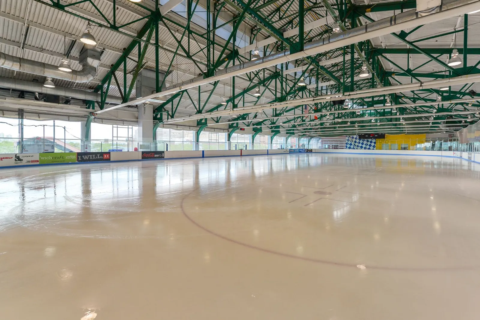 Empty ice sheet under the green industrial roof trusses of Pier 61, with sponsor boards (Coca-Cola Zero, Under Armour) along the dasher, floor-to-ceiling windows showing the Manhattan skyline, and a scoreboard and checkered wall mural at the far end. — Sky Rink at Chelsea Piers