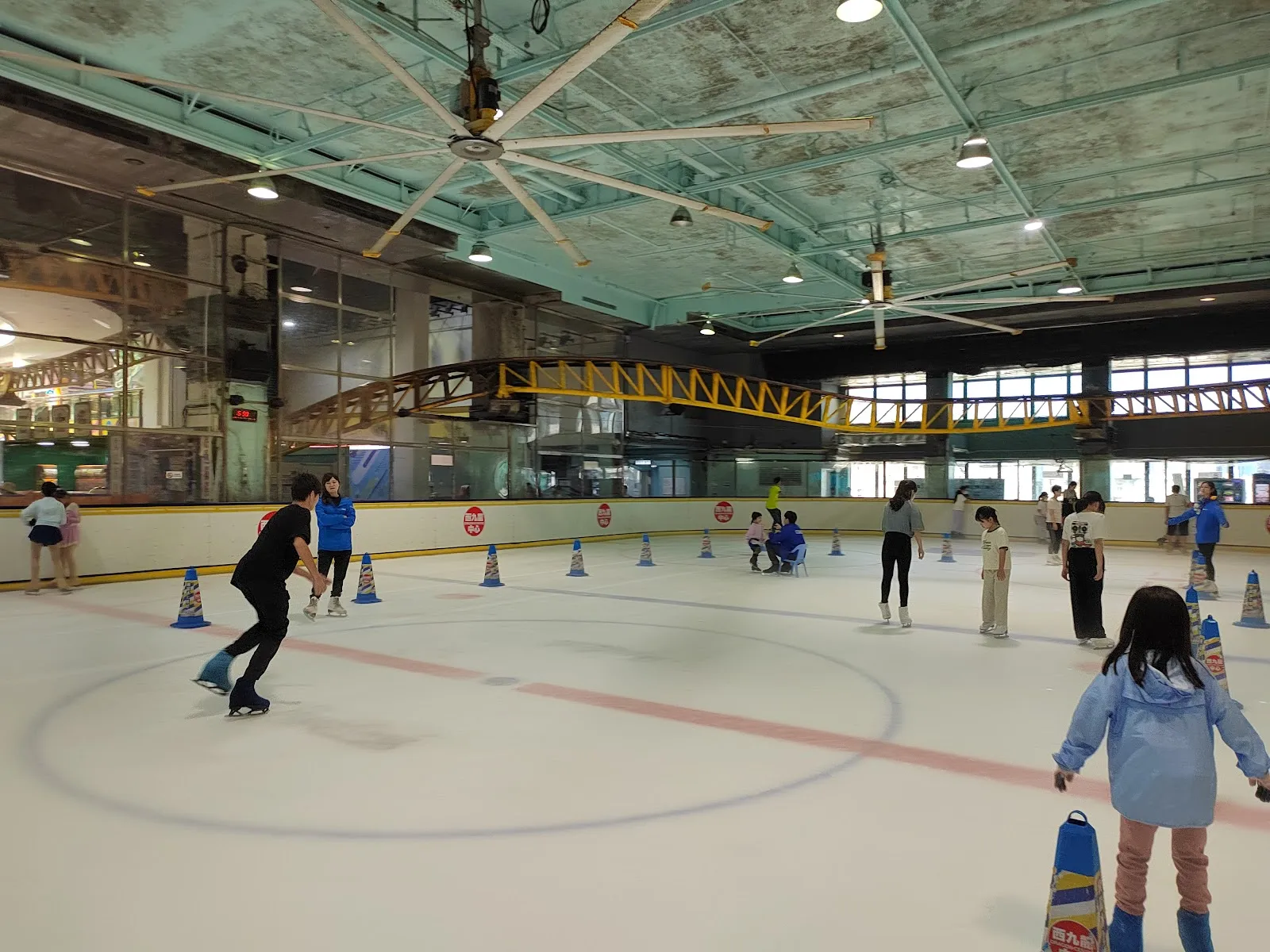 The Sky Rink ice with its centre circle and training cones under the roof structure of Dragon Centre, with a few skaters at the boards. — Sky Rink (Dragon Centre)
