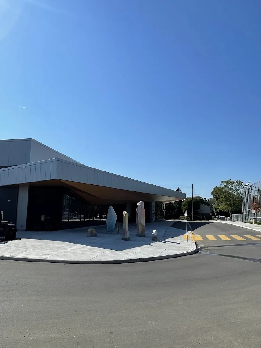 El nuevo edificio gris y bajo con un amplio techo en voladizo y fachada de vidrio, piezas de arte de piedra en la plaza y un paso peatonal en primer plano. — Skyway Community Centre