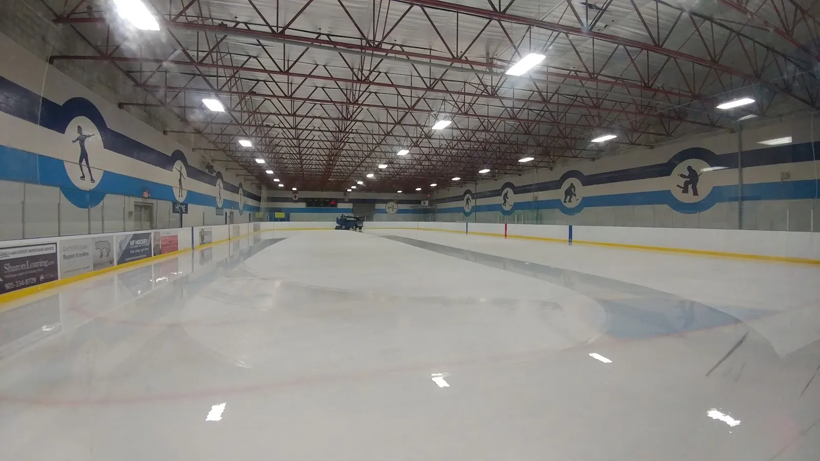 La nueva pista vacía desde una esquina: hielo brillante, vallas blancas con detalles azules y amarillos y una larga hilera de luces en el techo. — Skyway Community Centre