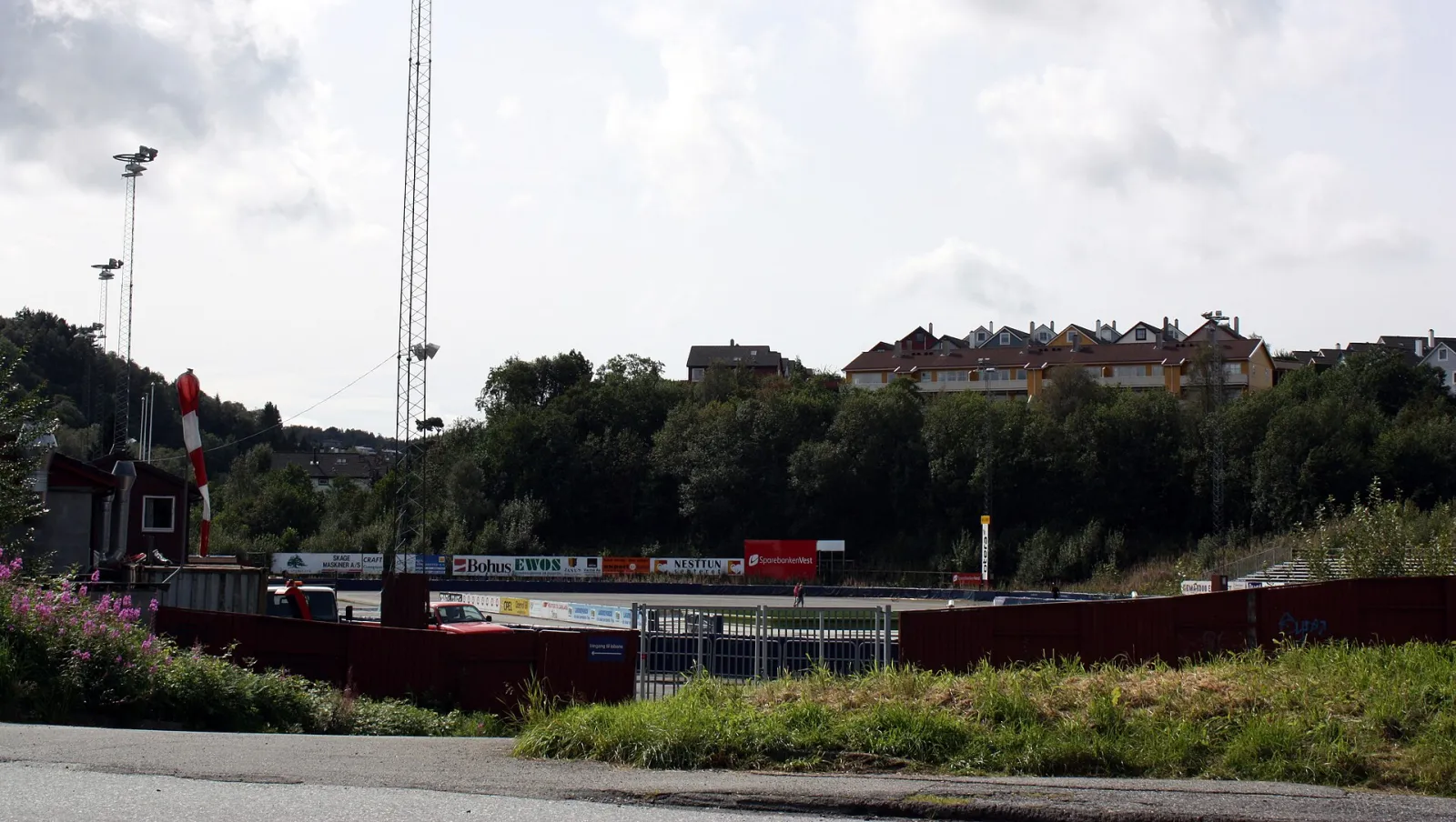 The Slåtthaug oval seen from the road in summer, behind its dark red boards and sponsor hoardings, with floodlight masts and the Fana hillside beyond. — Slåtthaug kunstisbane