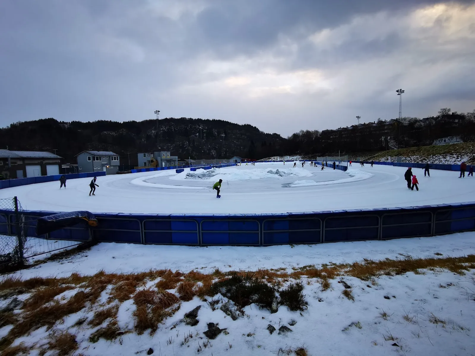 The blue-boarded Slåtthaug oval under a winter sky with a few skaters on the ice and snow along the infield. — Slåtthaug kunstisbane