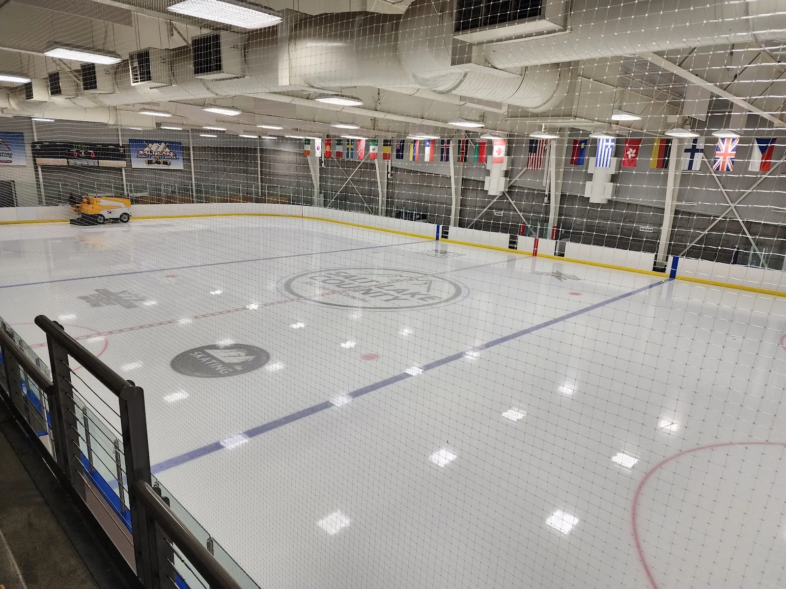 Una de las dos pistas vista a través de la red para espectadores, con un logotipo de Salt Lake County en el centro del hielo, una hilera de banderas nacionales en la pared del fondo y la pulidora de hielo estacionada en la esquina. — Salt Lake City Sports Complex