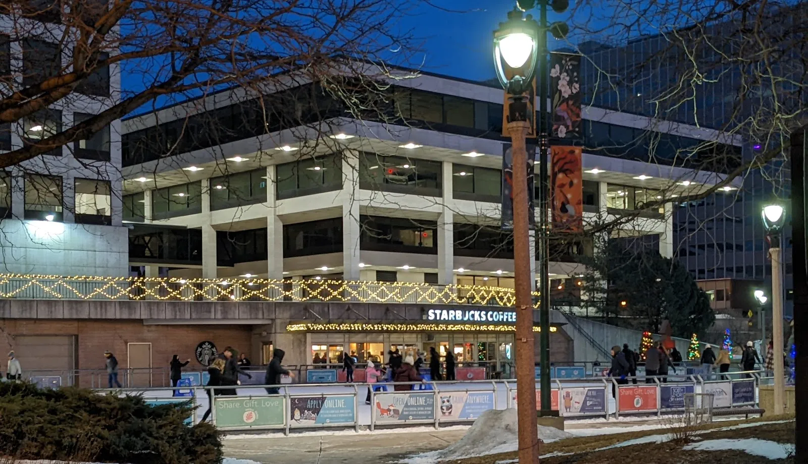 Skaters on the Slice of Ice rink at Red Arrow Park, Milwaukee, with the warming house and downtown buildings (Google Maps listing photo) — Slice of Ice at Red Arrow Park