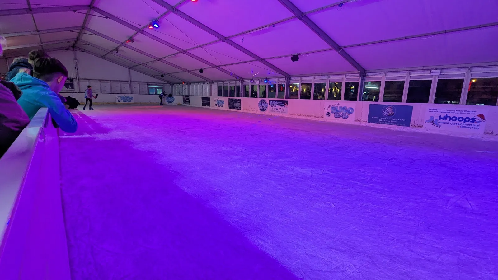 The tent-covered ice rink lit in purple, with the boards, sponsor banners along the far wall and the empty ice surface. — Sligo On Ice