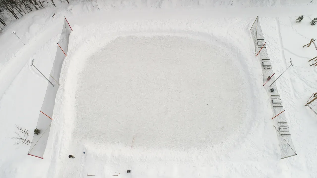 An aerial view of the rounded ice surface at Slora in winter, with the boards and netting at each end and tracks in the snow around it. — Slora kunstisbane (Lillestrøm)