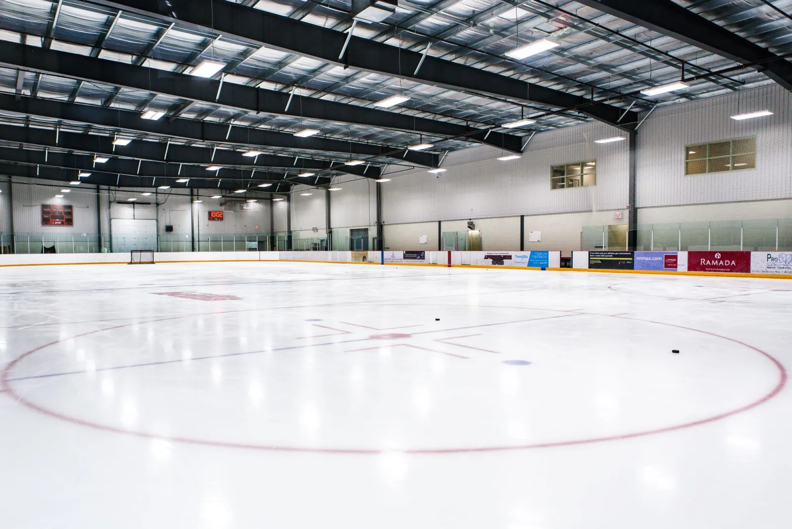 Una pista vacía de tamaño NHL recién inundada vista desde el centro de la pista, con las armaduras de acero del techo y anuncios de patrocinadores a lo largo del lado opuesto. — SLS Centre