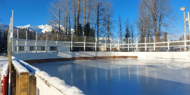 The outdoor rink in Smithers, a sheet of ice ringed by wooden boards and tall bare trees, with snow-covered mountains behind. — Smithers Outdoor Rink
