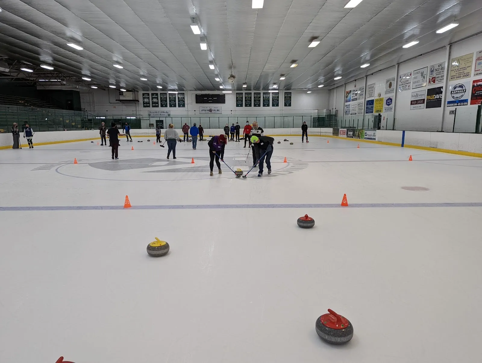 Una sesión de curling en la pista, con jugadores y piedras repartidos por el hielo entre conos naranjas bajo un techo claro con una franja de ventanas y banderines en el extremo opuesto. — Smithfield Municipal Ice Rink