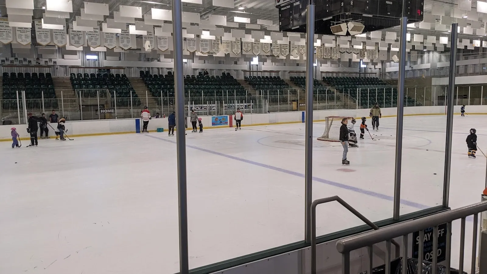 A wide view of the sheet through the glass, players spread across the ice under a high ceiling with green seating along the far side. — Smiths Falls Memorial Community Centre