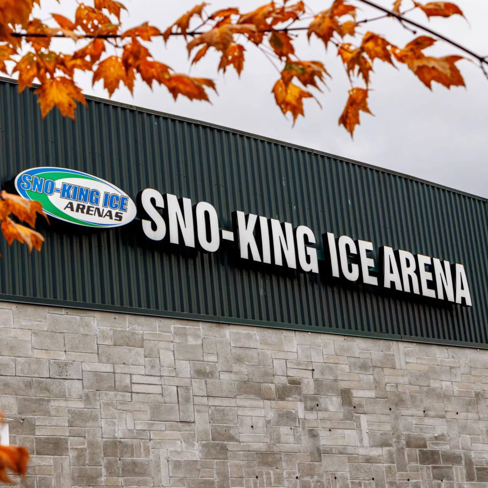 Close-up of the building's stone-veneer wall and dark corrugated-metal upper facade with the illuminated "SNO-KING ICE ARENA" sign and oval logo, autumn maple leaves in the foreground. — Sno-King Ice Arenas - Renton