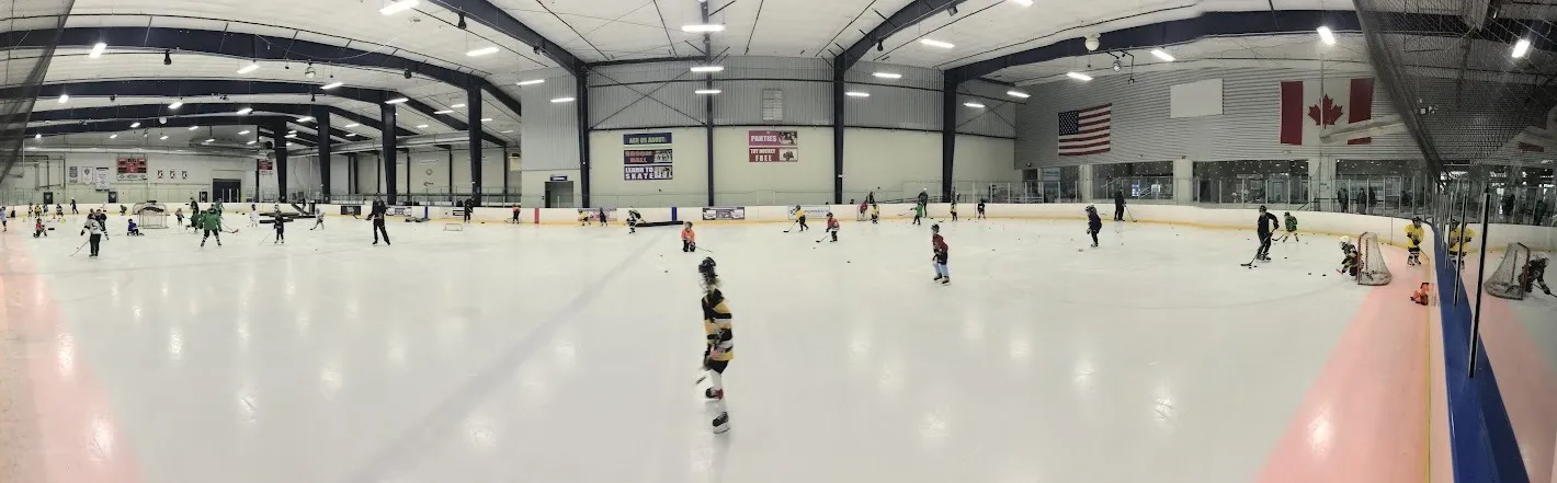 A panorama of a Sno-King Renton sheet during a youth practice, under navy steel trusses with the US and Canadian flags on the end wall — Sno-King Ice Arenas - Renton