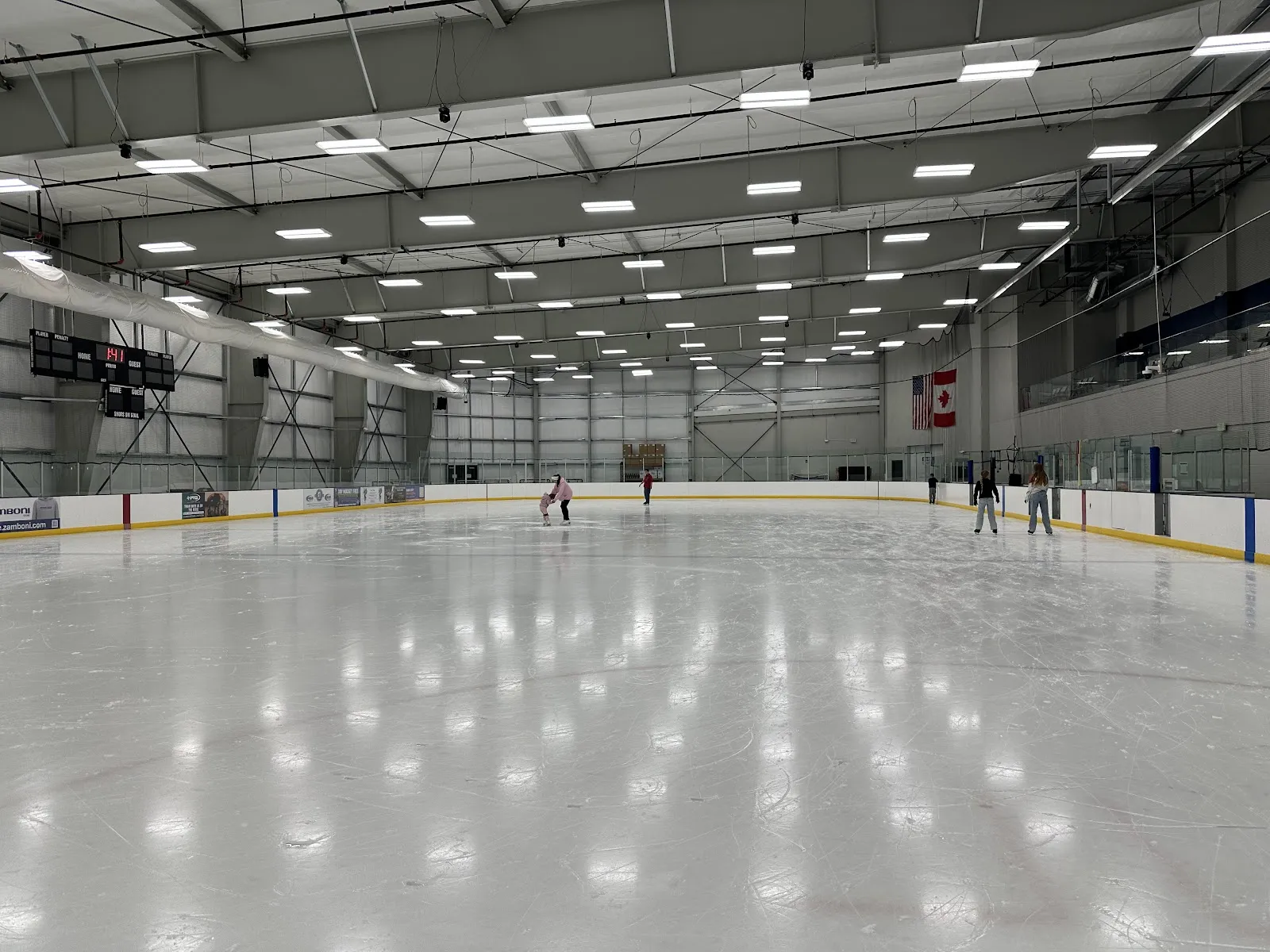 Amplia vista interior de la pista a nivel del hielo, con toda la pista, las vallas, la iluminación superior, el marcador y banderas colgadas de Estados Unidos y Canadá, con algunos patinadores a lo lejos. — Sno-King Ice Arenas - Snoqualmie