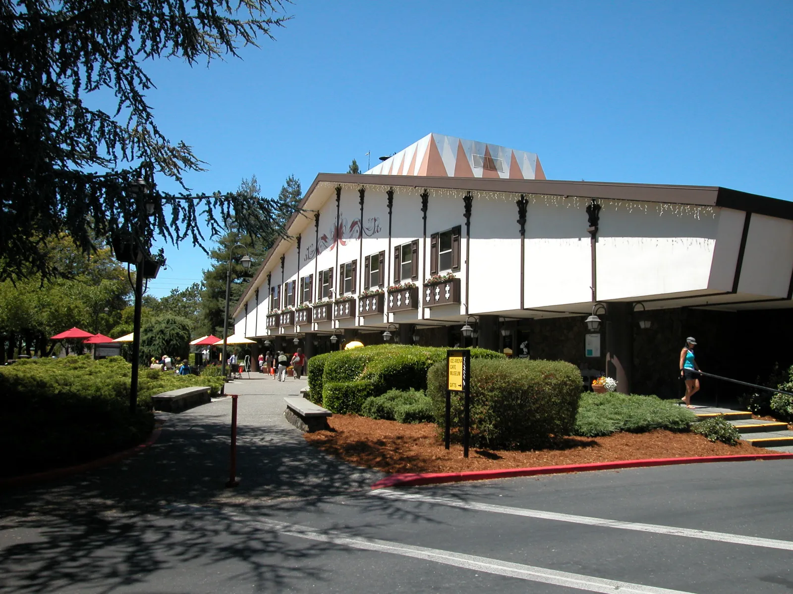 Two-story white building with dark chalet-style trim and a peaked, diamond-patterned roof; a walkway leads to an entrance with a sign reading 'ICE ARENA CAFE MUSEUM GIFTS', trees and red patio umbrellas nearby. — Snoopy’s Home Ice