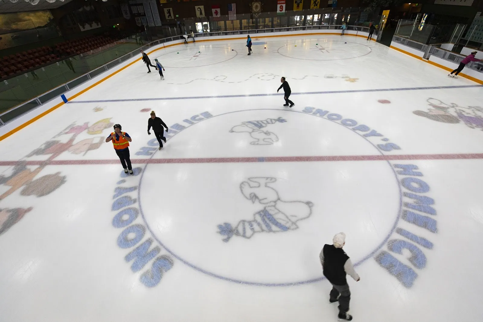 The sheet seen from above with the Snoopy's Home Ice name and logo at centre, a few skaters and players around it. — Snoopy’s Home Ice